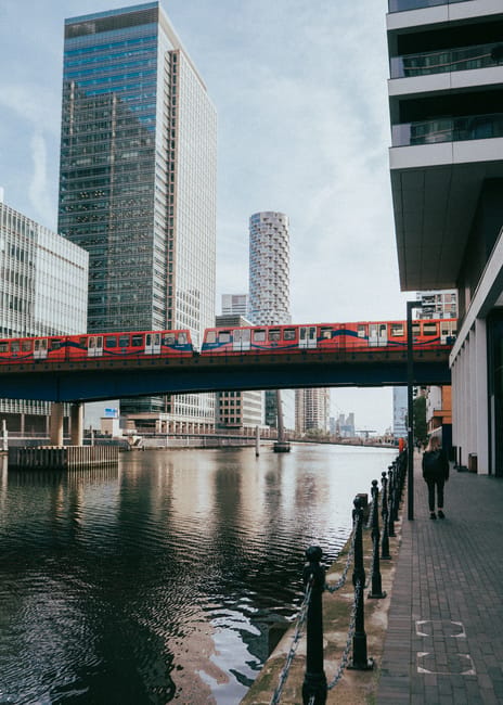 A panoramic view of Canary Wharf in London featuring a modern city skyline with tall skyscrapers made of glass and steel, with reflective surfaces shining under a clear blue sky. In the foreground, a 