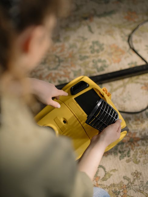 A person holding a yellow portable carpet cleaning machine with a black handle and vents, positioned on a patterned area rug in a room with exposed brick wall in the background. The individual’s han