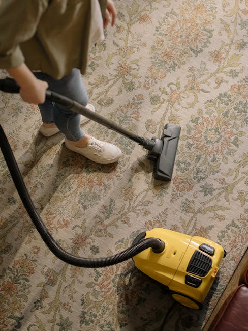 A person holding a yellow portable carpet cleaning machine with a black handle and vents, positioned on a patterned area rug in a room with exposed brick wall in the background. The individual’s han