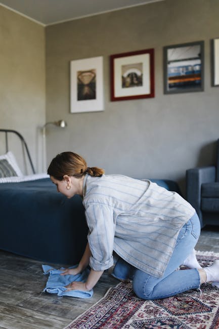 A senior man with white hair and a beard is vacuuming a yellow and white patterned area rug in a bright living room, while a young boy with red hair sits on the edge of a beige sofa with multicolored 
